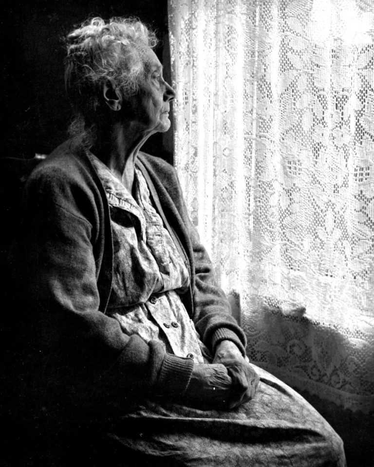 Black and white image of a woman sitting by a window looking through a lace curtain