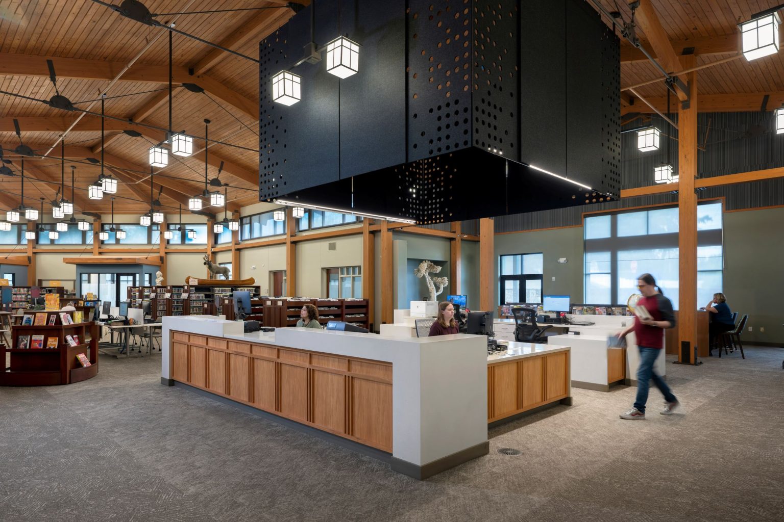 Woman sitting at circulation desk inside a library. A patron walks by the desk.